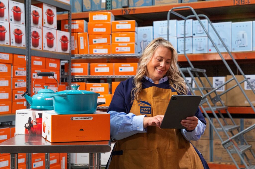 A woman in a warehouse uses a tablet while standing beside Le Creuset cookware boxes and kitchen items on a metal table. Shelves filled with orange boxes are in the background.